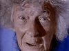 Close-up of an elderly person with wide eyes and gray frizzy hair in a blue garment, staring at the camera with a startled look—evoking the suspenseful style of the “Don’t Look in the Basement (1973) DVD.”.