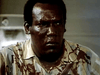 A man with a serious expression, his shirt and face splattered with blood, stands indoors under harsh lighting—evoking classic scenes from the Don’t Look in the Basement (1973) DVD.
