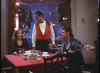 A waiter in a red vest stands between a blonde woman and a dark-haired man at a restaurant table with cups, a Keno sign, and the Final Impact (1992) DVD. The softly lit room features plants as décor.