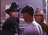 A man in a black cowboy hat and denim jacket stands beside a woman in a white hat with a pink band, both looking at another man, evoking the atmosphere of classic films like Final Impact (1992) DVD.