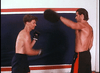 Two shirtless men are boxing indoors, training with focus pads like a scene from Final Impact (1992) DVD. Both wear boxing gloves and shorts, channeling the energy of a classic kickboxing movie.