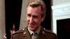 A man in a brown military uniform with light hair smiles indoors, standing before a fireplace and dark curtains—reminiscent of scenes from Attack on the Iron Coast (1968) DVD, a rare classic movie.