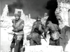 Four soldiers in heavy gear move through ruins with smoke swirling behind them, echoing German paratrooper film classics. This scene captures the intense wartime atmosphere of "The Green Devils of Monte Cassino (1958) DVD.