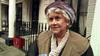 An older woman with curly gray hair, a mauve hat, and beige scarf stands outdoors by black railings and a cream building—evoking scenes from rare DVDs like Mrs. Palfrey at the Claremont (2005) DVD.