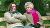A young man in a chunky sweater and scarf chats with an older woman in a beige coat, pink scarf, and black gloves on a park bench about the "Mrs. Palfrey at the Claremont (2005) DVD" as trees sway gently behind them.