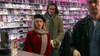 A woman in a red coat and scarf, a man with a banana, and another man in a leather jacket stand at the counter of a DVD store lined with rare movies, including "Mrs. Palfrey at the Claremont (2005)" on DVD, displayed behind them.