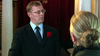 A man in a suit with a red flower in his lapel stands indoors, looking seriously at a blonde woman seen from behind. Dark red curtains and columns evoke the intrigue found in the Mrs. Palfrey at the Claremont (2005) DVD.