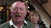 An older mustached man speaks with his mouth open as an older woman with short hair stands behind him, looking surprised. They are indoors near a staircase—a scene from the "Mrs. Palfrey at the Claremont (2005) DVD.