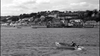A person rows a small boat across the water, with a coastal town and hillside homes behind—a scene reminiscent of classic films like Murder Ahoy (1964) DVD featuring Miss Marple.