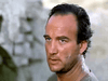 A man with short dark hair and a serious look stands outdoors, his face sweaty and dirty in rocky terrain—recalling classic films. Inspired by scenes from Sahara (1995) DVD.