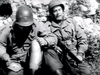 Two soldiers in helmets and uniforms sit against a stone wall; one holds a rifle, the other bandages his leg. This black-and-white image is from "Then There Were Three" (1961) DVD, capturing the realism of vintage classic films.