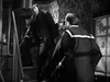 Three men in dark clothing stand on a narrow staircase in a dimly lit, vintage room—evoking the suspenseful atmosphere of classic war films like "Went the Day Well? (1942)" on DVD.