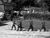 Six soldiers in uniform march in formation along a sunlit path near a hedge, with a quaint house and gravestones behind—evoking scenes reminiscent of classic war films like Went the Day Well? (1942) DVD.