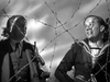 Two men stand behind barbed wire, looking anxiously skyward—one in a suit with binoculars, the other in a navy uniform. The tense scene evokes classic war films like Went the Day Well? (1942) DVD.