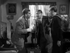 Three men—one in a suit, one in a sailor uniform, and one in a cardigan—converse tensely indoors near floral décor and a staircase, capturing the suspenseful mood of classic war films like Went the Day Well? (1942) DVD.
