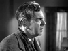 Black and white profile of an older man with wavy, graying hair in a houndstooth jacket and scarf, looking deep in thought—evoking a character from "Went the Day Well? (1942)" DVD. A blurred curtain and wall fade into the background.