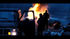 A silhouetted person stands near an open car door at dusk, while another faces a flaming vehicle—an intense scene reminiscent of the suspense found in Killshot (2008) DVD. Debris and a parked car are visible in the foreground.