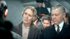 An older woman with short gray hair stands next to an older man with a mustache, both looking concerned in a tense scene from the "A Is For Acid (2002) DVD," with people and a uniformed officer blurred in the background.