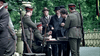 Uniformed soldiers sit and stand around an outdoor table, checking documents of two civilians. The scene recalls moments from the historical film "Einstein and Eddington (2008) DVD," with trees and a decorative fence in the background.