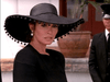 A woman dressed in black with a stylish hat stands outdoors, capturing the mystique of hard-to-find films like Trouble in Paradise (1989) DVD, while a suited man lingers near a potted plant in the background.