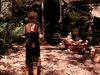 A woman in a black dress stands outside as a man in a wooden tub wearing a party hat excitedly waves his arms—like a scene from "Trouble in Paradise (1989)" DVD—set in lush tropics near a rustic cabin.
