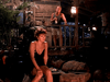 A woman in a black dress sits pensively on a crate as a man with a bottle stands on a deck behind her, surrounded by tropical plants. The dim light and rustic mood evoke the intrigue of rare films like the Trouble in Paradise (1989) DVD.