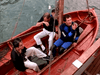 Four men in a small wooden sailboat—one in a wetsuit, others casually dressed—look up with concern, recalling scenes from classic films like Trouble in Paradise (1989) on DVD.