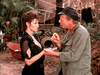 A woman in a black dress holds hands with a man in a green jacket and black cap, outdoors in a rustic setting—evoking classic scenes from the Trouble in Paradise (1989) DVD.