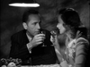 A man and woman share wine at a small table under a hanging lamp in a dimly lit, intimate setting—evoking the classic romance of "Passage to Marseille (1944)" DVD. A bowl of fruit sits beside them.