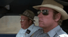 Two uniformed police officers sit side by side in a car, reminiscent of a scene from the "White Lightning (1973) DVD." The officer on the right drives wearing sunglasses, while his partner on the left looks straight ahead.