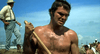 A shirtless man, sweating and gripping a shovel under a bright sky, evokes the gritty tension of "White Lightning (1973)"—now available on DVD. In the background, another man in a hat and suspenders watches intently.