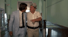 Two men stride down a hallway, deep in conversation. One wears a light blue suit and tie; the other sports a white shirt, glasses, hat, and items on his belt—reminiscent of characters from the White Lightning (1973) DVD.