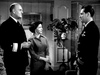 A man in a naval officer’s uniform faces another man in uniform and a woman in a dress, in a room with a plant and framed pictures—evoking classic scenes from "Submarine Command (1951)" on DVD.