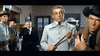 Five men stand indoors in a tense scene from Three Graves for a Winchester (1966) DVD. One, in a light suit, holds a riding crop, while another in a black hat and brown coat points a pistol. Wallpaper and a door form the backdrop.