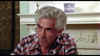 An older man with gray hair and a serious expression sits by a white ornate fireplace, wearing a red, white, and blue plaid shirt. A Dead of Night (1974) DVD rests on the mantel beside a blue vase and brass lamp.