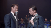 Two men in tuxedos stand at microphones on stage, evoking iconic duo Dean Martin and Jerry Lewis, as a band plays in the background—scene from the "Martin & Lewis (2002) DVD.