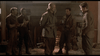 Five men in WWII uniforms talk in a dimly lit room with straw and rustic furniture, like a scene from the Ardennes forest. One holds a mug as another faces the group. Available on A Midnight Clear (1992) DVD.