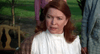 A woman with long auburn hair, resembling Ellen Burstyn from Resurrection, sits outdoors in a white high-collared blouse and brown checked shawl, looking concerned—evoking the feel of the Resurrection (1980) DVD.