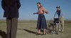 A woman tentatively walks on grass away from a wheelchair beneath a clear sky, recalling the stirring near-death scenes in Resurrection (1980) DVD. A man holds the wheelchair as another person stands close by.