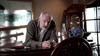 An older man with gray hair and a beard leans on a table, holding a glass of whiskey. Nearby rest a bottle, blue flowers, and the "Blue-eyed Butcher (2012) DVD" case, with a framed picture and cabinet in the background.