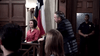 A woman in a red shirt sits in the witness stand as a man in a suit stands nearby, with a large flag and police officer present—a gripping scene from Blue-eyed Butcher (2012) DVD, perfect for true crime and rare movie collectors.