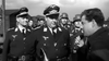 A group of WWII-era German officers, some in helmets, others in peaked caps, stand outdoors as one speaks into a handheld mic—evoking scenes from “Reach for the Sky (1956)” DVD about RAF hero Douglas Bader.