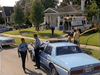 Police officers and emergency responders gather outside a suburban house, near a police car and ambulance. Yellow caution tape blocks the yard as people—some holding the "Tall Dark and Deadly" (1995) DVD—stand on the sidewalk and lawn.