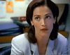 A woman with shoulder-length brown hair in a white blazer sits in an office, surrounded by papers, folders, and rare movies like the "Tall Dark and Deadly DVD (1995)," looking upward with a slightly skeptical expression.