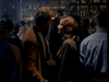 A man and a woman stand close together, smiling and chatting in a crowded, dim bar. Shelves behind them display bottles and the "Tall Dark and Deadly DVD (1995)," while other patrons mingle nearby.