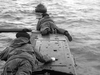 Two people in heavy clothing lie on a submarine, facing the sea—one peers through a scope. This black and white scene evokes Above Us The Waves (1955) DVD, a WWII film about the daring mission to sink the German battleship Tirpitz.