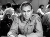 A man in military uniform sits at a table, looking serious, evoking the intensity of an aerial warfare briefing in "The Wild Blue Yonder (1951) DVD," with others talking in the blurred black-and-white background.