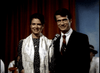 A woman in a patterned dress and scarf stands beside a man in a dark suit with a red striped tie. Smiling at the mic on stage with red curtains, they could be presenting the "Murder By Reason of Insanity (1985) DVD.