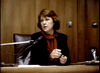 A woman with shoulder-length brown hair sits at a desk in a wood-paneled room, dressed in a dark blazer and red blouse, reflecting the tense mood of "Murder By Reason of Insanity (1985) DVD," with a microphone, papers, and coffee pot on the desk.