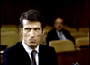 A man in a suit and tie sits in a nearly empty courtroom, looking to his right with a serious expression—evoking the suspense of Murder By Reason of Insanity (1985) DVD. Another person is seated in the out-of-focus background.